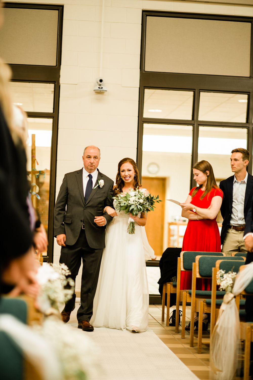 Father of the bride in dark suit escorts smiling bride in white gown carrying greenery bouquet down aisle.