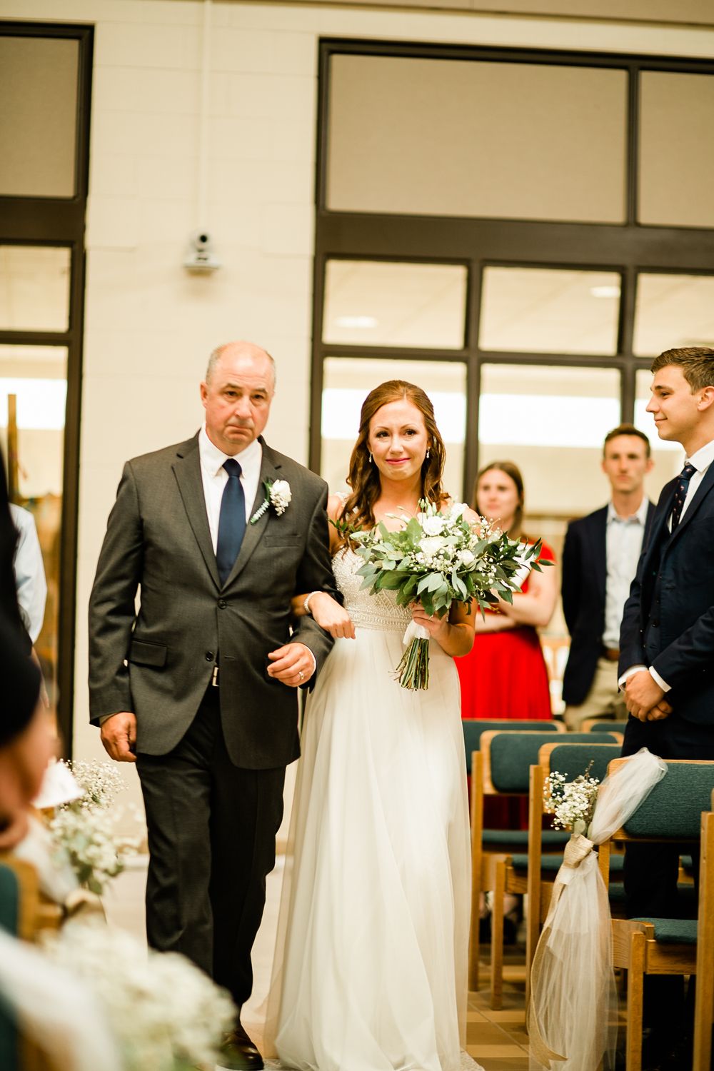 Father walks radiant bride in white wedding gown down church aisle as guests stand and watch ceremony.