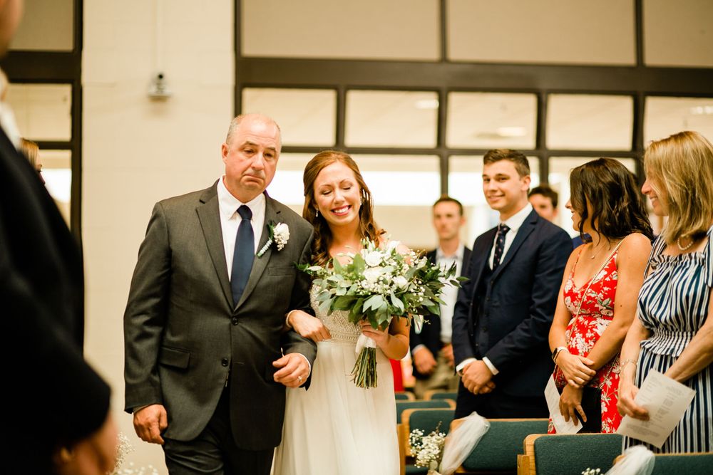 Proud father in dark suit escorts smiling bride in white dress carrying white floral bouquet down aisle.