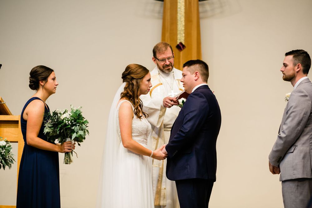 Officiant performs wedding ring exchange ceremony as bride in white gown and groom in navy suit hold hands.
