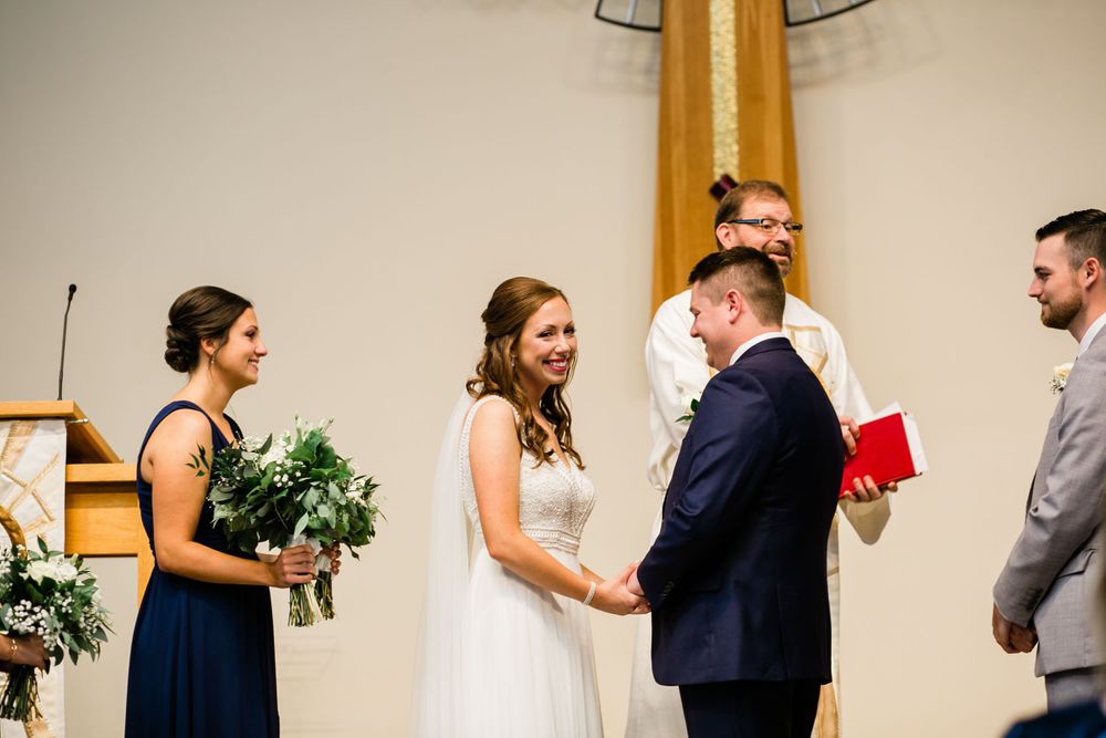 Smiling bride and groom hold hands at the altar with officiant and bridesmaid during wedding ceremony.