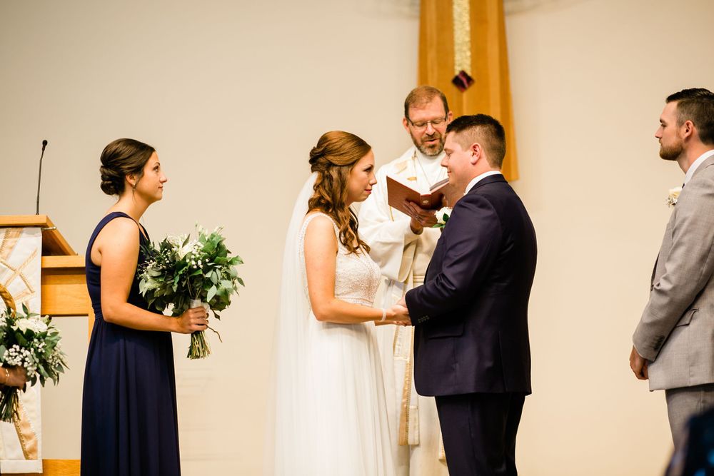 Bride and groom exchange vows at church altar with officiant and wedding party during marriage ceremony.