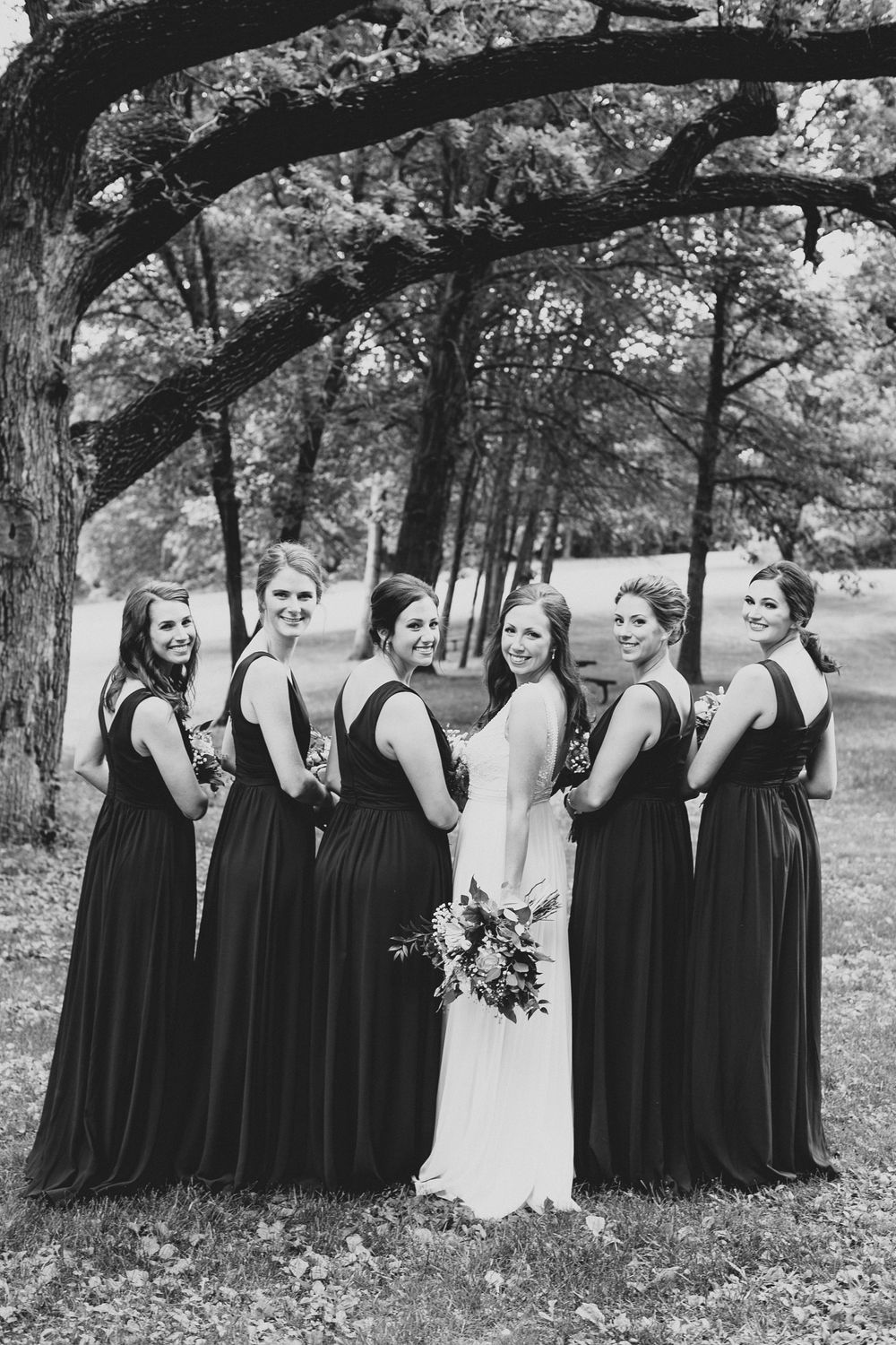 Black and white photo of bride with bridesmaids in long dark gowns posing under large tree branches.