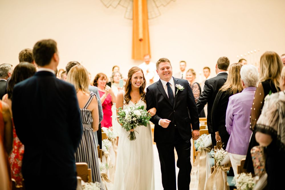 Bride walks down the church aisle with escort past seated wedding guests holding a white bouquet.