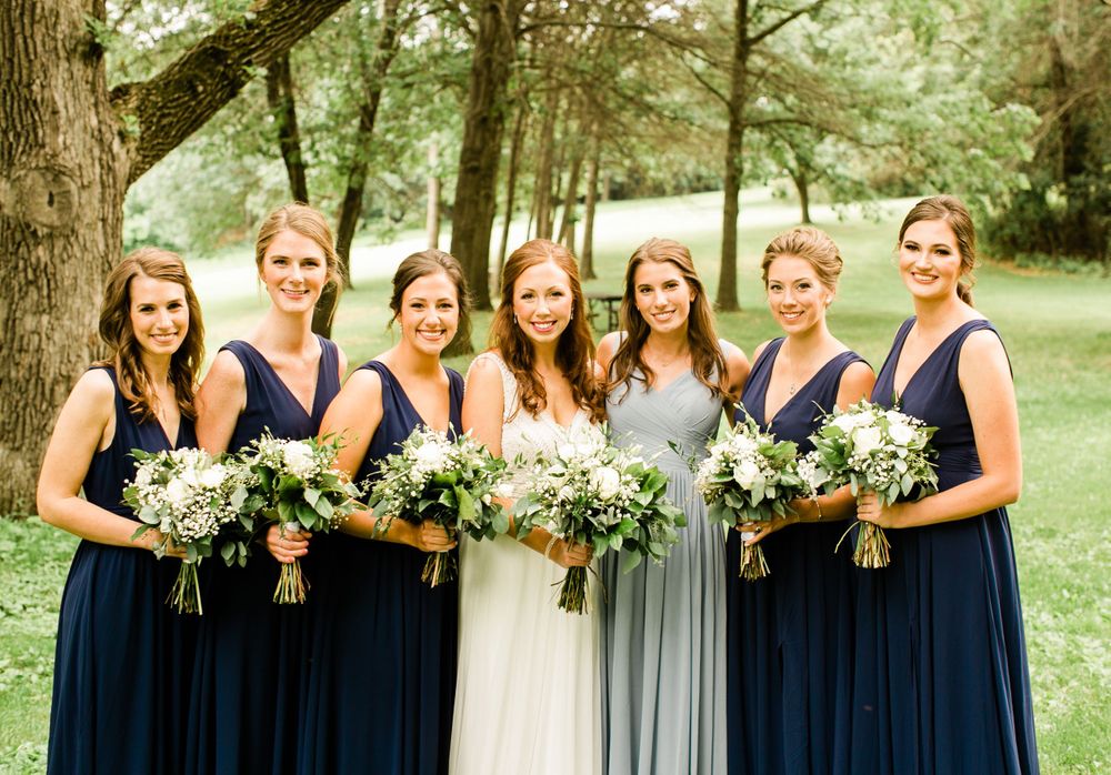 Bride with six bridesmaids in navy and gray gowns holding white floral bouquets on lush green lawn.