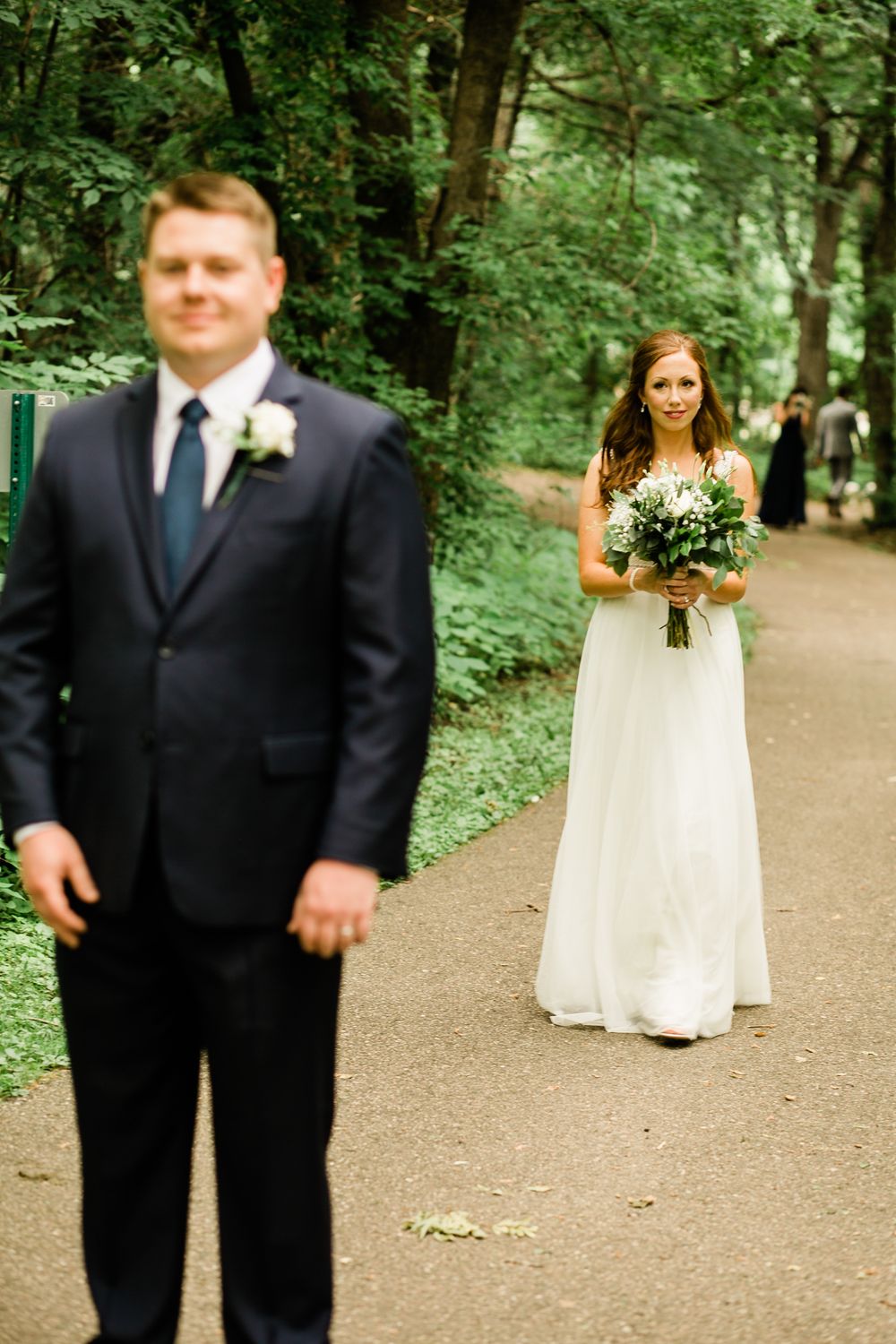 Bride in white gown approaching groom on tree-lined path for first look.