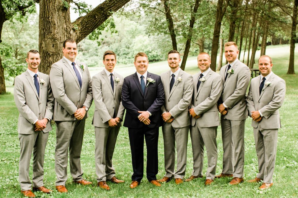 Groom in navy suit stands with eight groomsmen in gray suits and navy ties on green lawn.