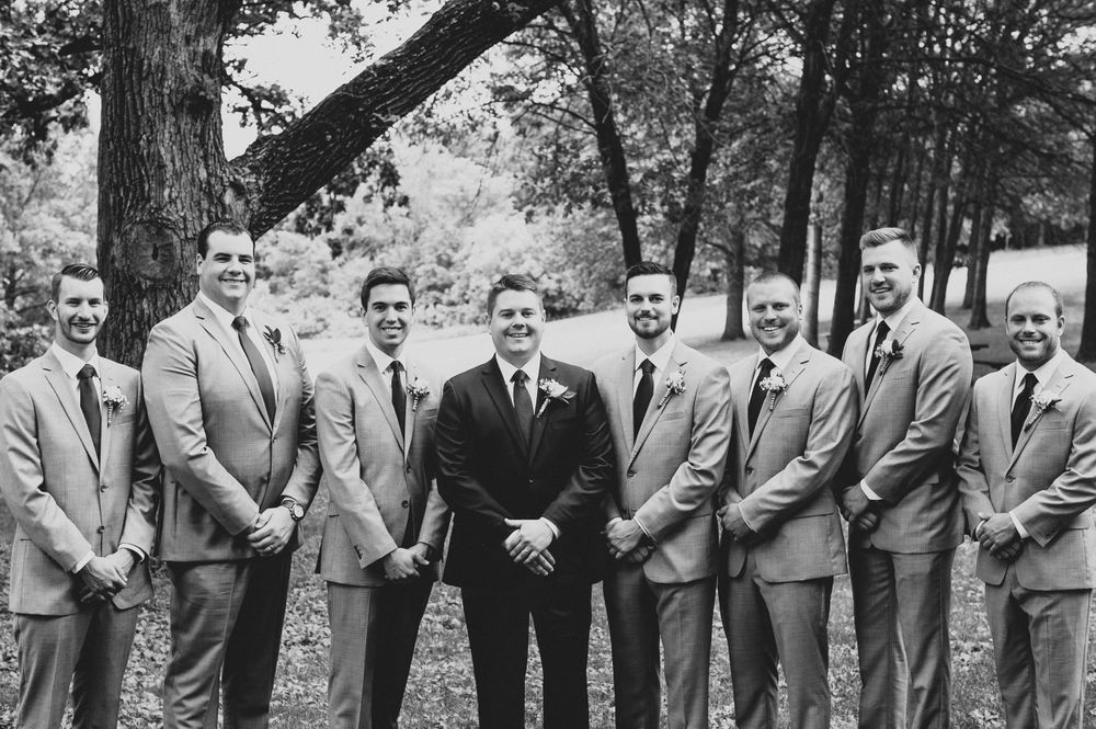 Black and white photo of groom in dark suit flanked by eight groomsmen in light suits outdoors.