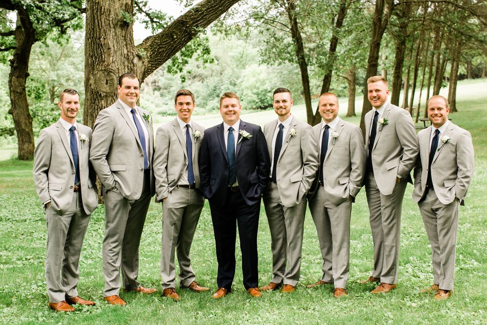 Groom in navy suit smiles with eight groomsmen wearing gray suits and navy ties under large trees.