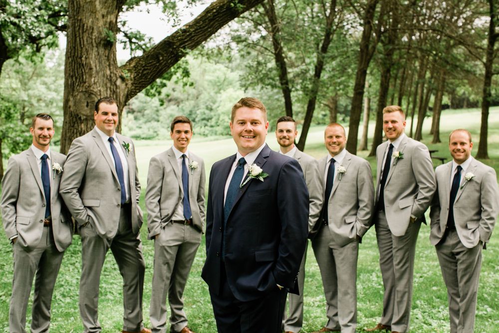 Groom in navy suit stands in front of eight groomsmen in gray suits arranged on green grass.