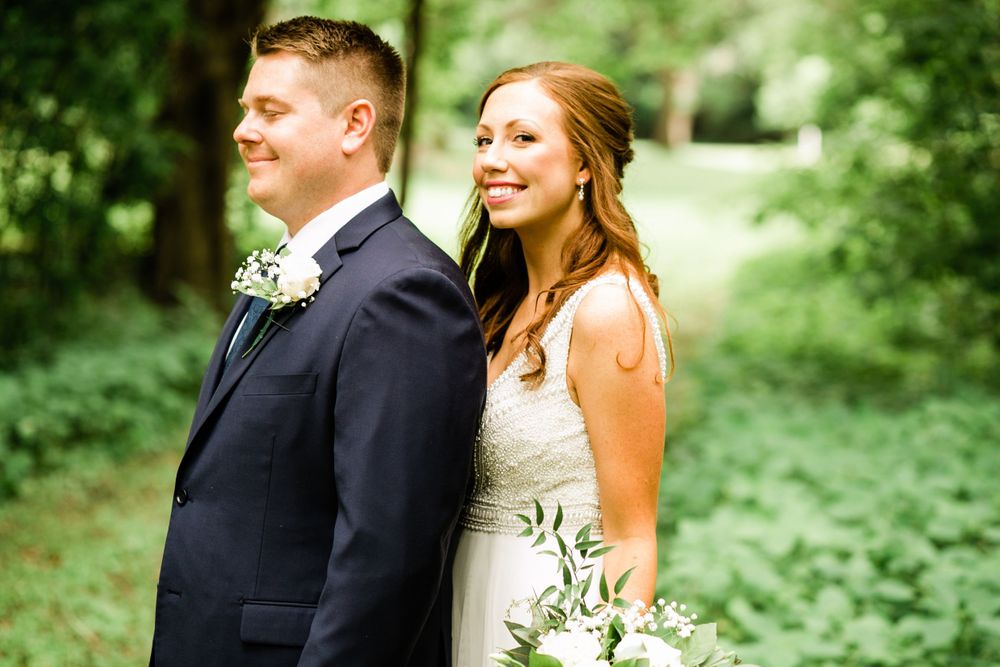 Bride and groom stand back-to-back smiling in lush green outdoor setting.