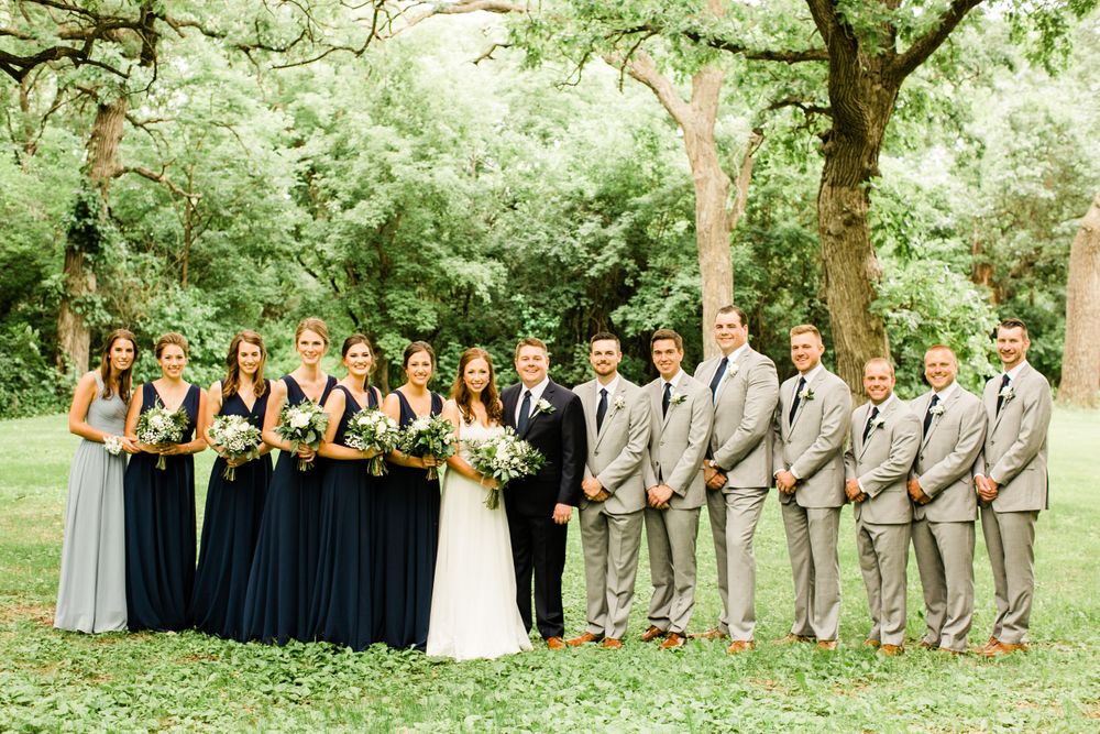 Wedding party posing outdoors with bridesmaids in navy gowns and groomsmen in gray suits among lush green trees.