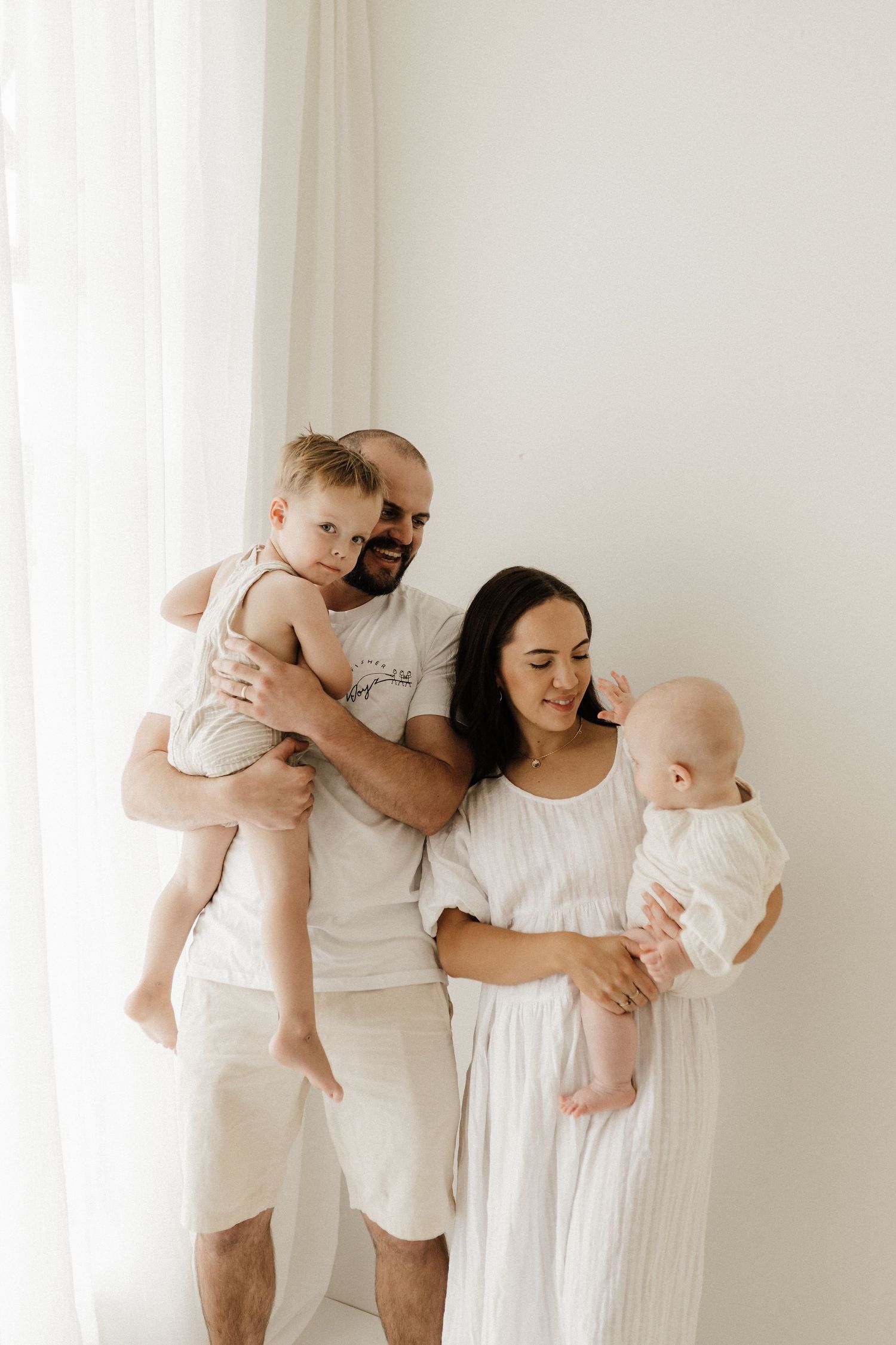 Family in white clothing holding two babies while standing by bright window.