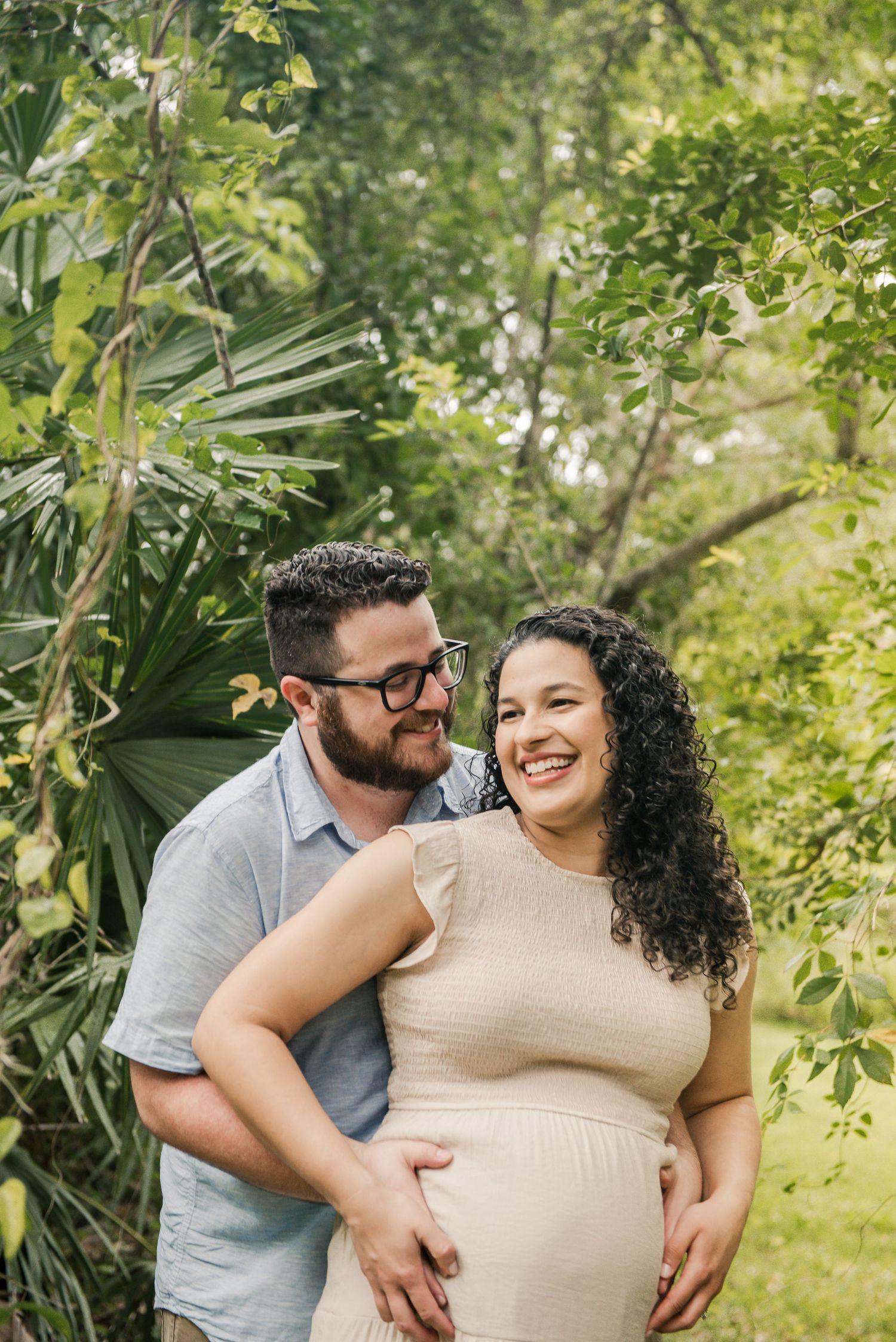 A couple embraces in a lush garden setting during a maternity photoshoot amid green foliage and natural sunlight.