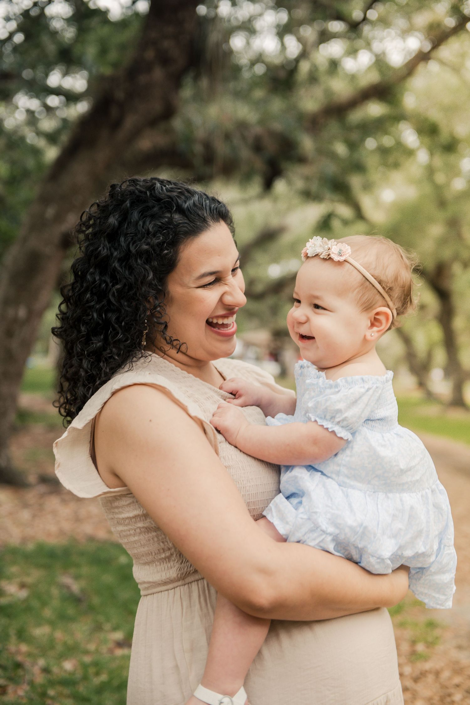 A candid outdoor portrait of a woman in a beige dress sharing a joyful moment with a baby in white lace dress.