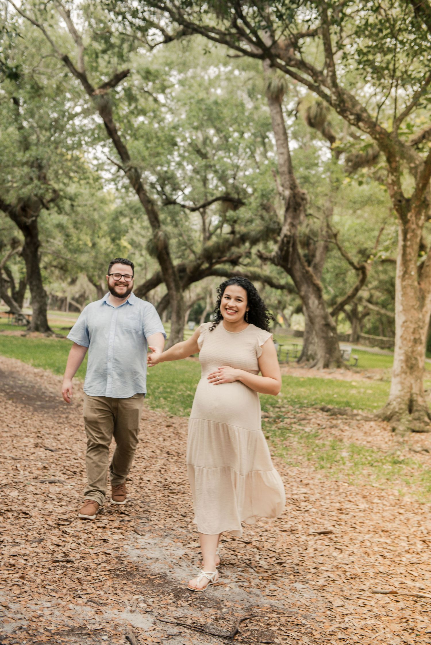 A couple walks together on a leaf-covered trail through a wooded park on a sunny afternoon.