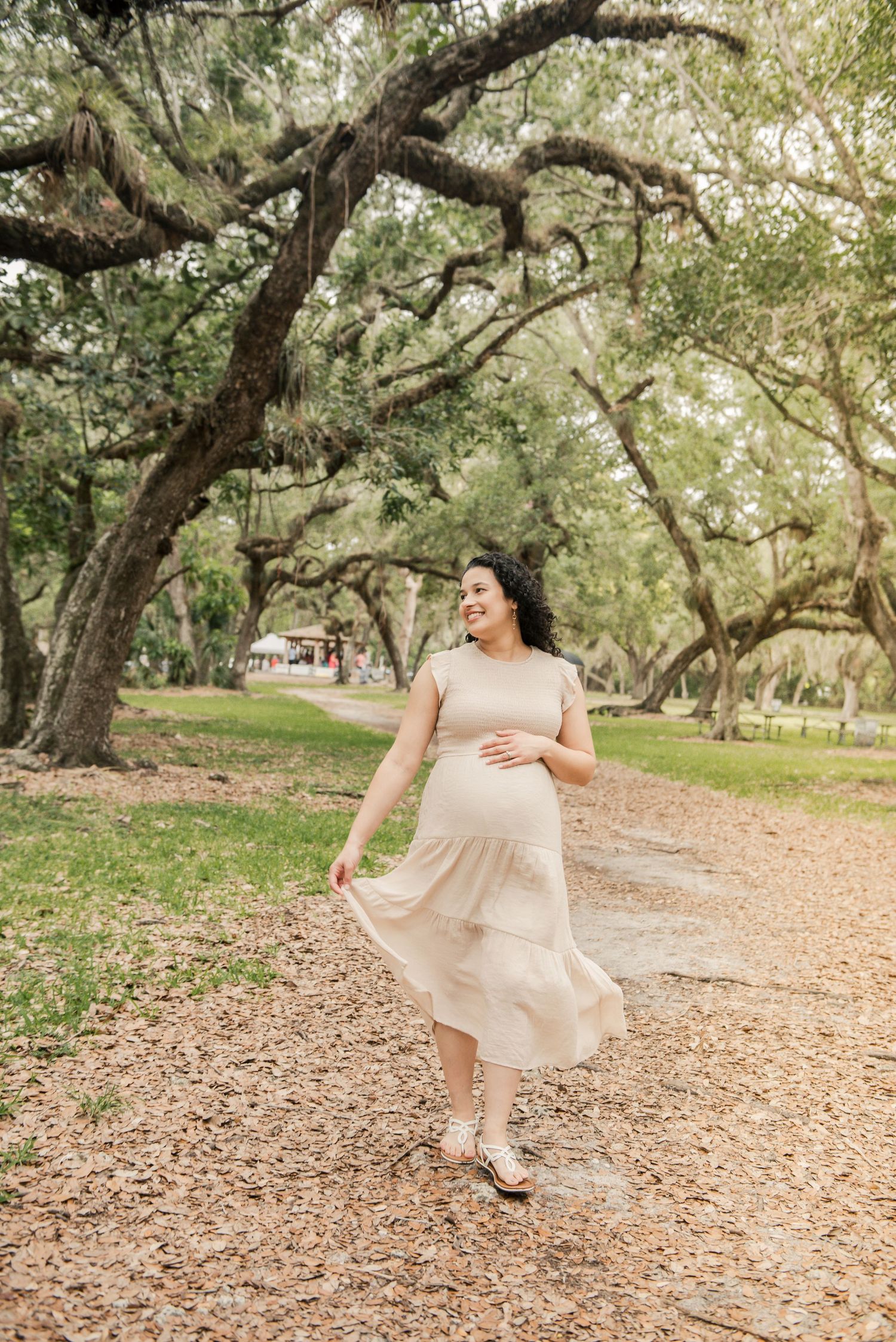 An expectant woman in a flowing beige dress twirls on a path beneath large oak trees draped in Spanish moss.