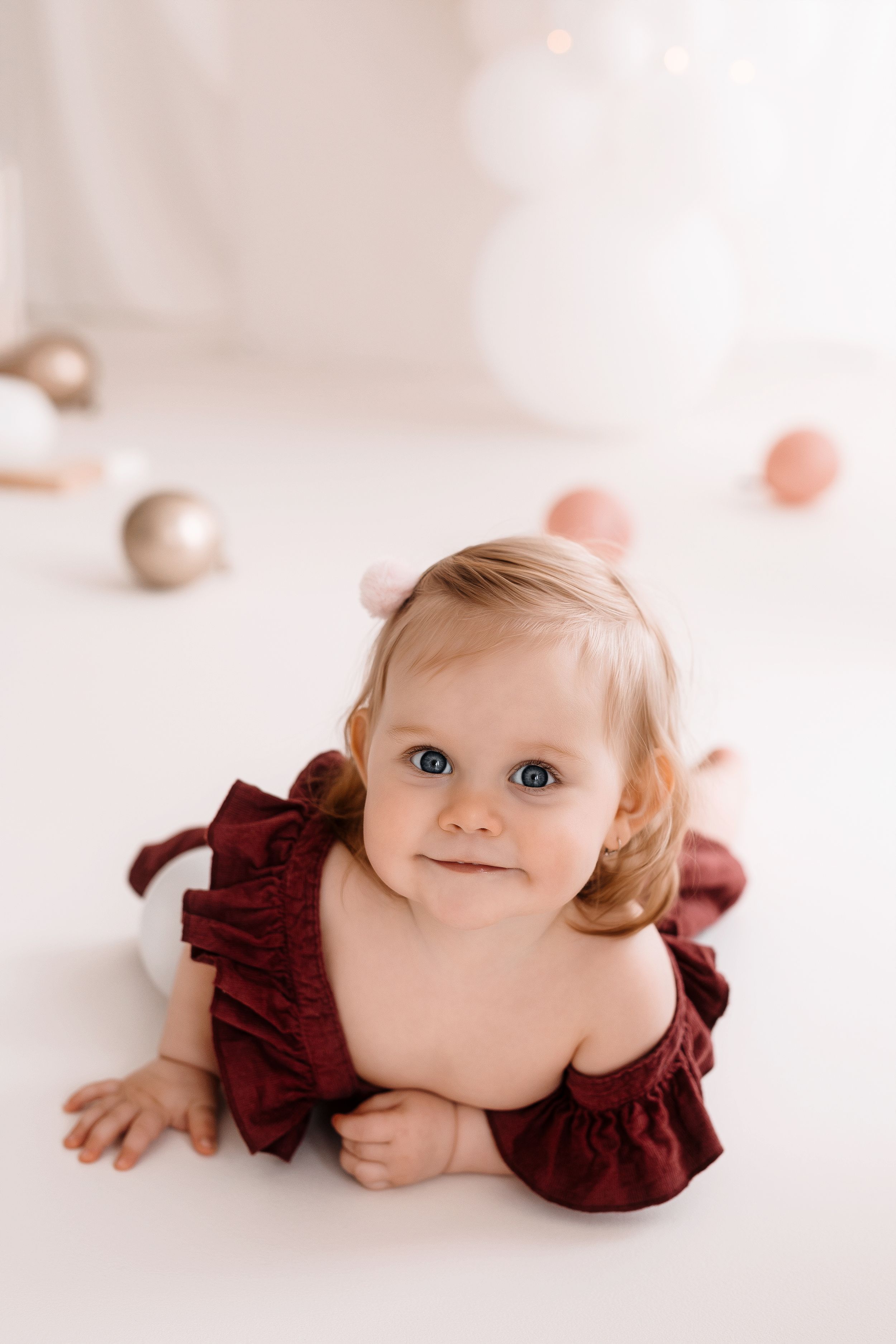 A toddler in a burgundy ruffled outfit lies on a white floor with gold ornaments in a soft studio setting.