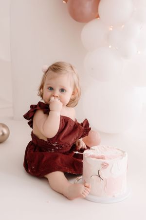 A toddler in burgundy dress reaching for a small pink and white birthday cake.