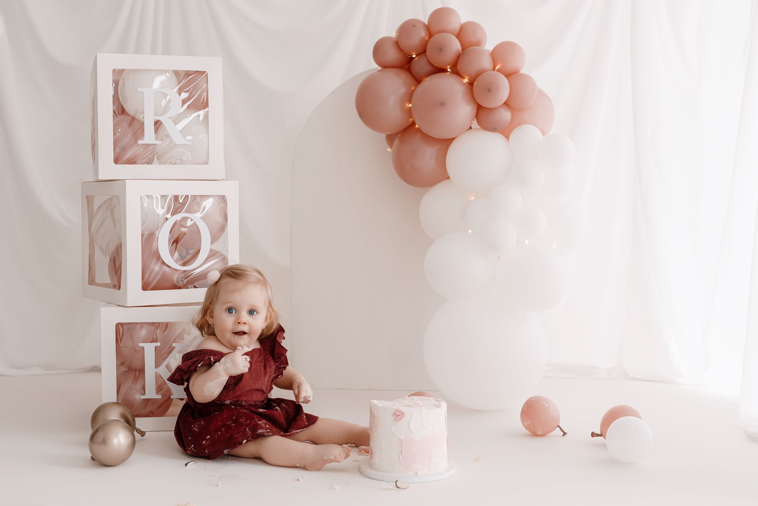 Vintage style first birthday photo setup with pink balloons and letter blocks against white backdrop.