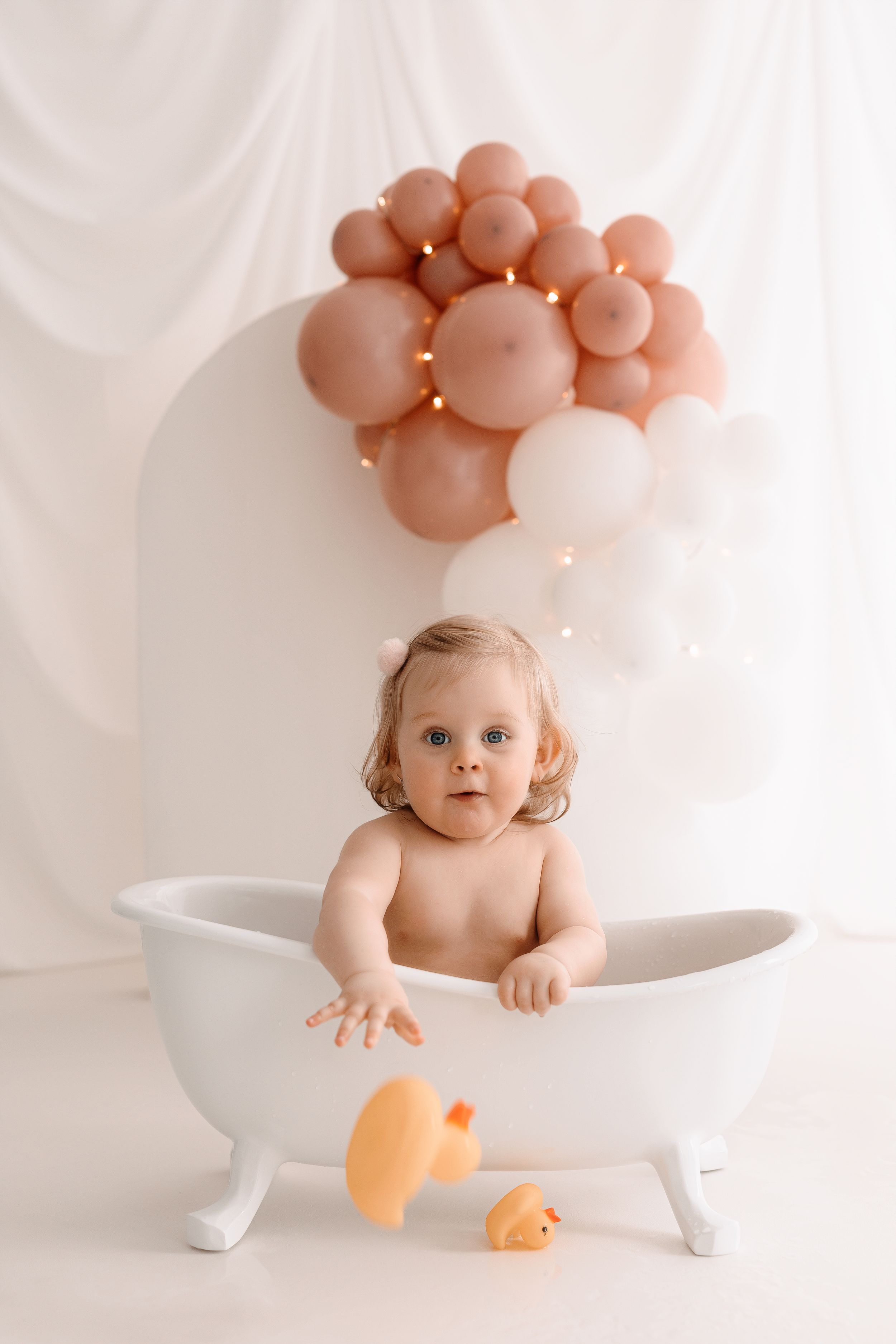 A charming bathtub photo setup with rose gold balloons and rubber ducks.