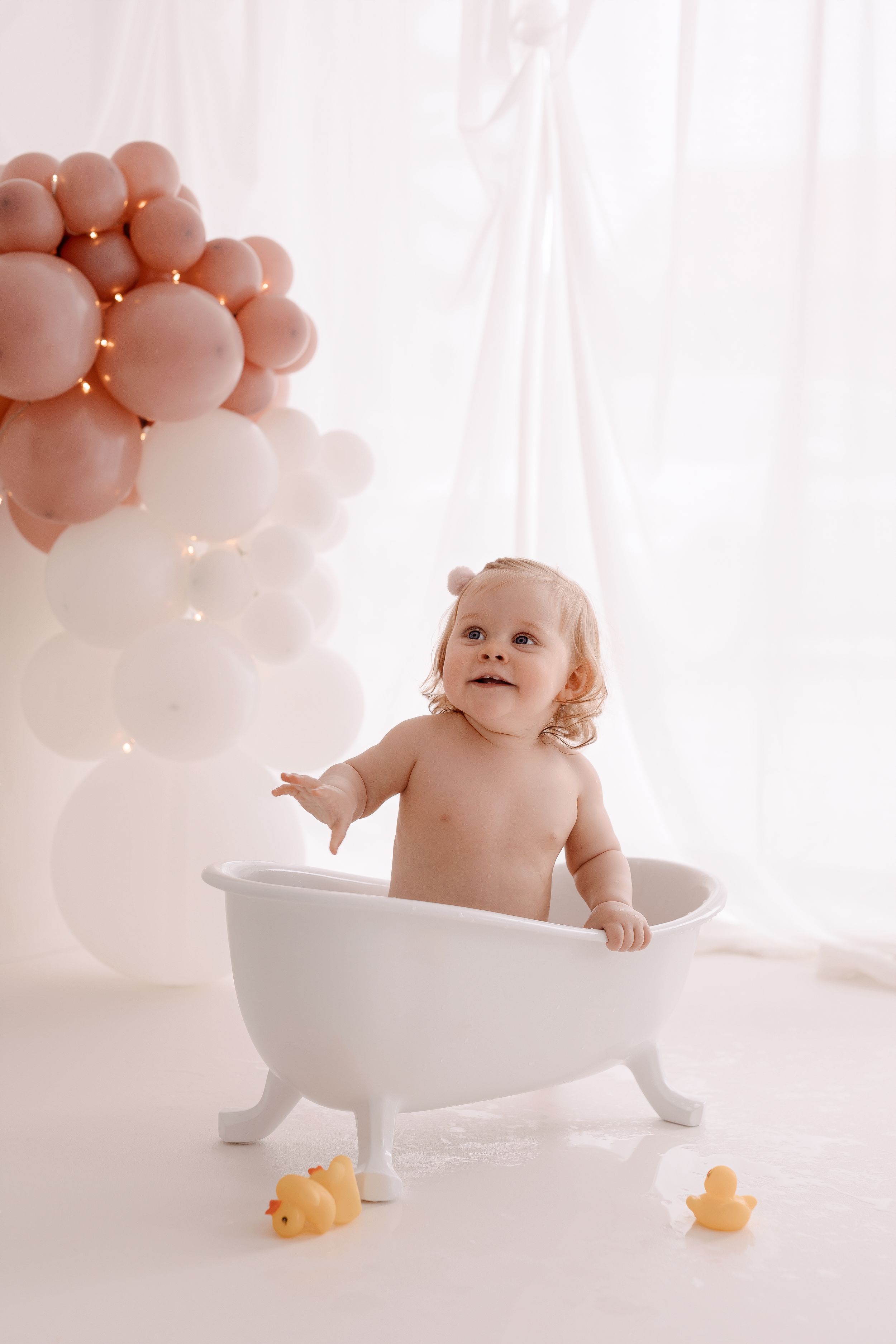 A toddler standing in a white clawfoot tub with rose gold balloons and rubber ducks.