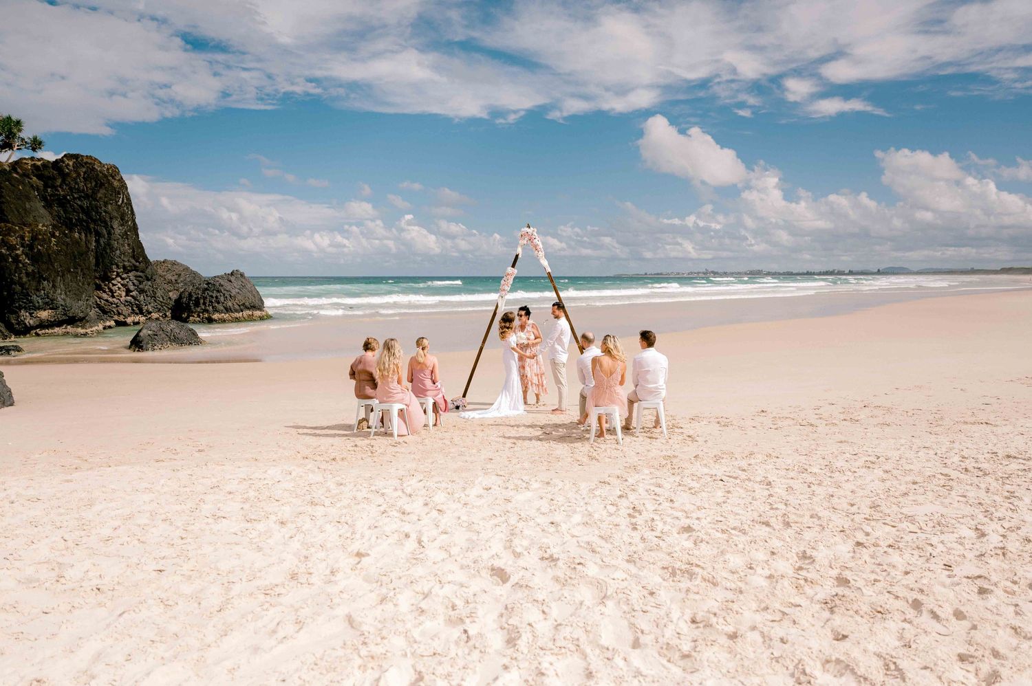 Beach wedding ceremony with white arch and pink-clad wedding party on sandy shore.