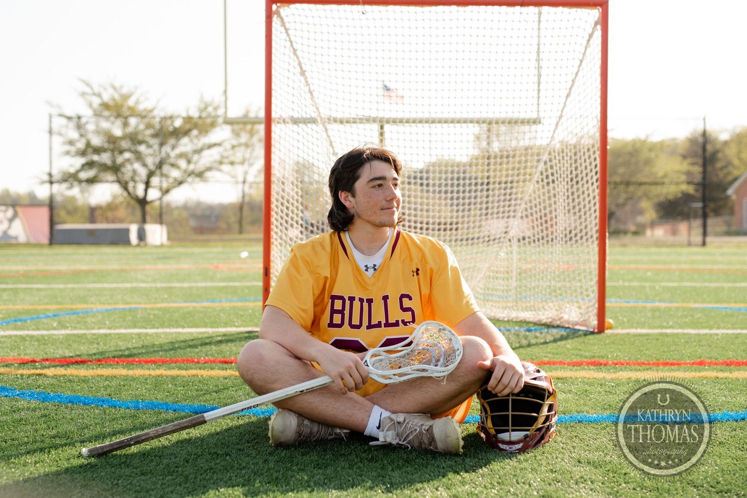 Player in Bulls uniform sitting cross-legged with lacrosse gear by goal net.