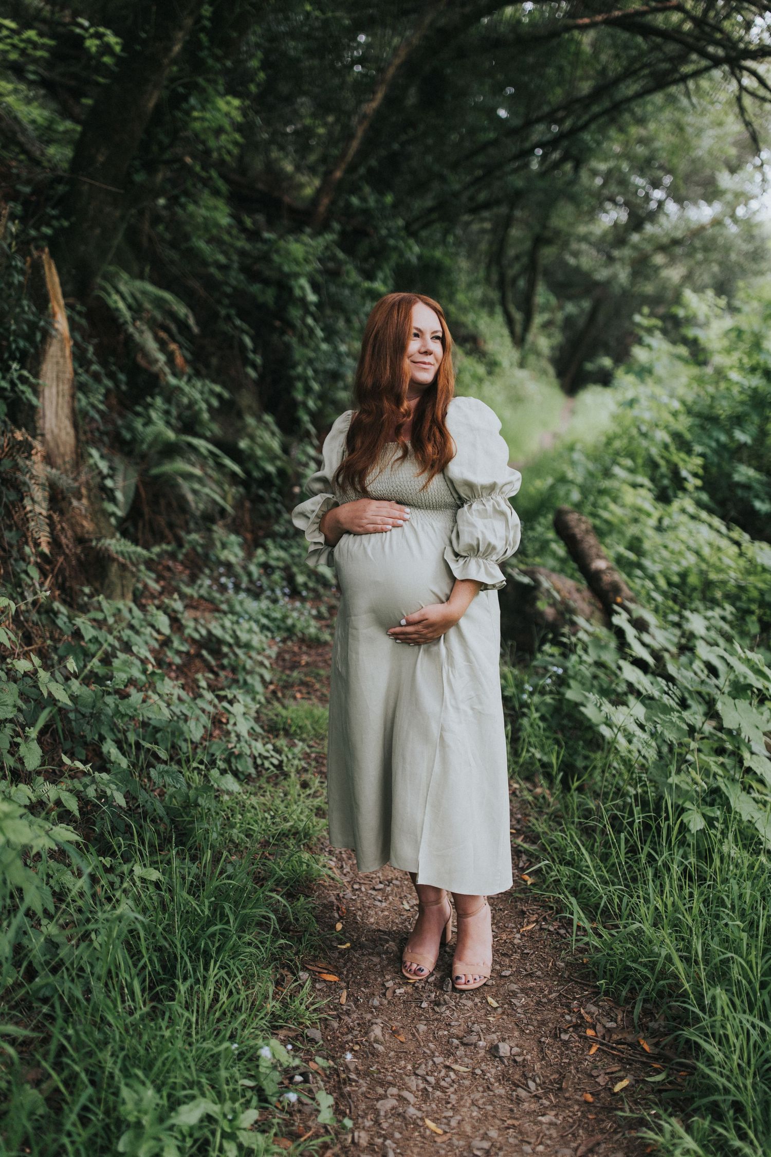 Expectant mother in a flowing white dress stands on a woodland path surrounded by greenery.