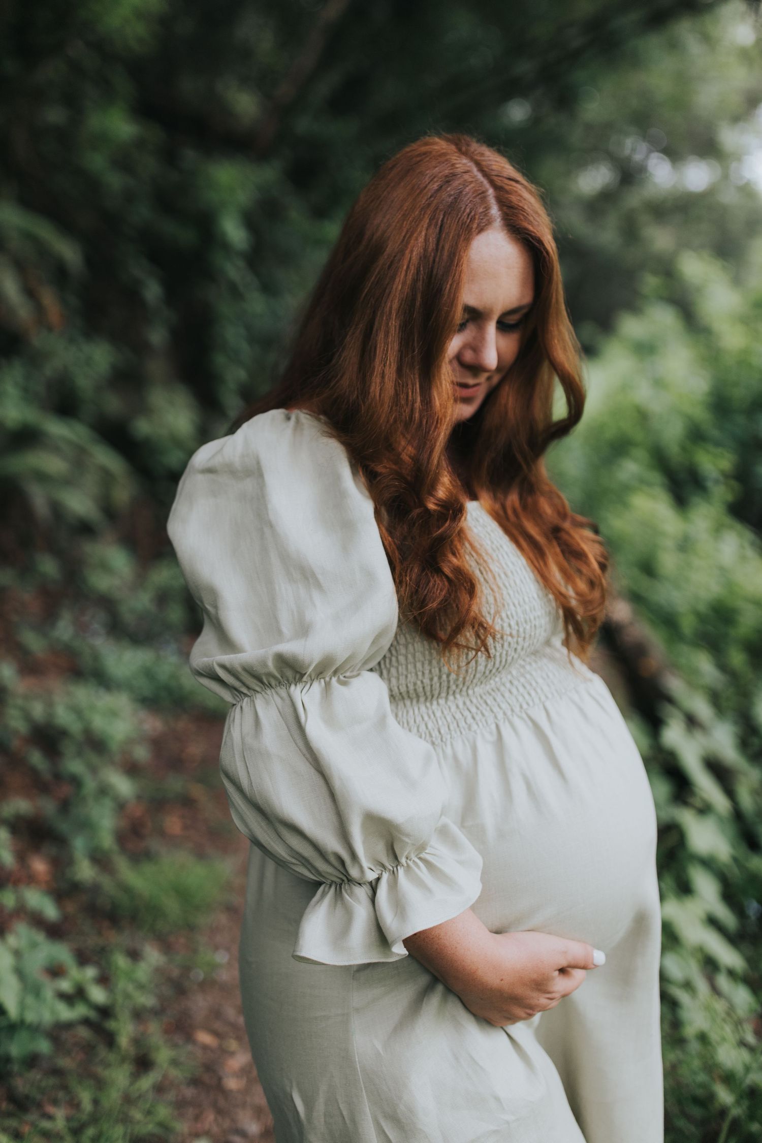 A maternity photoshoot in nature featuring an elegant white dress with puff sleeves against a lush green backdrop.