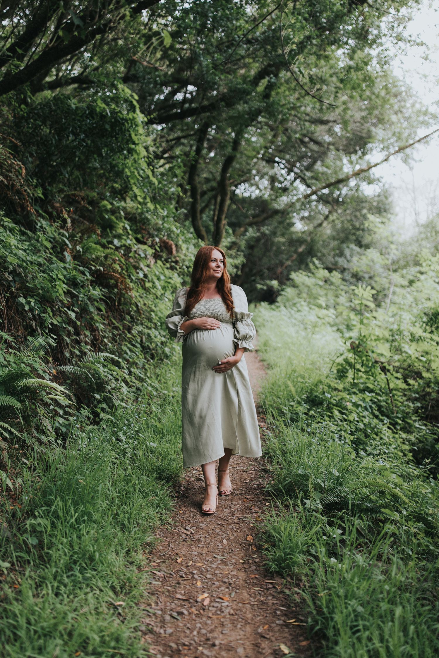 Barefoot expectant mother walks along a wooded trail wearing a light-colored dress with puffy sleeves.