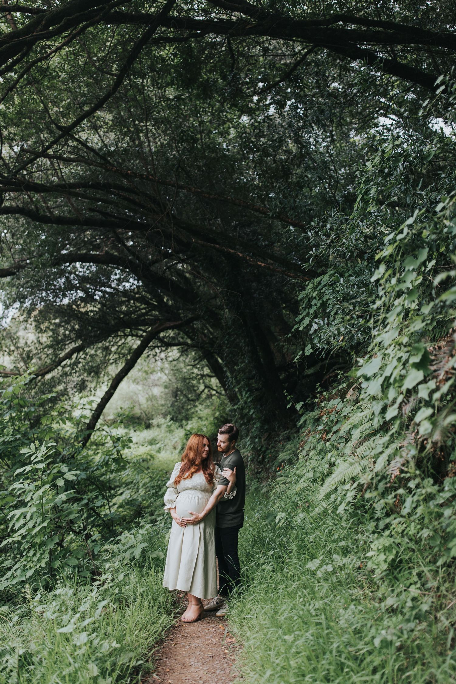 A couple shares an intimate moment under a natural tree archway during their maternity photoshoot.