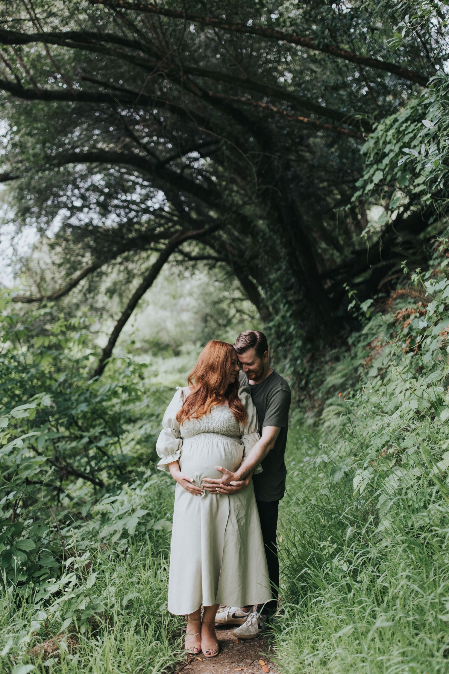 Intimate maternity photo of a couple on a wooded trail with overhanging branches and lush foliage.