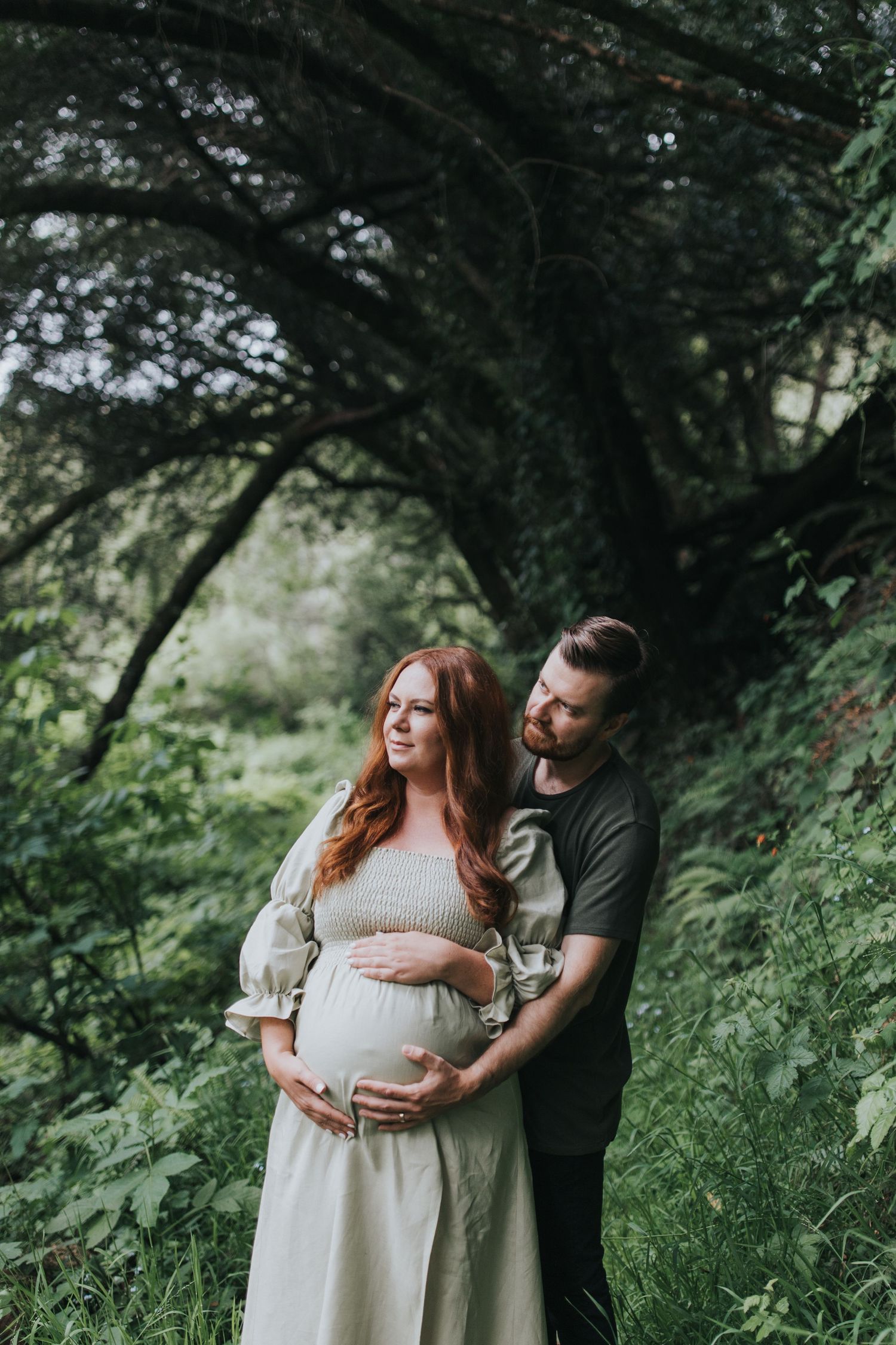 Maternity portrait of a couple embracing on a forest path with dramatic tree canopy overhead.