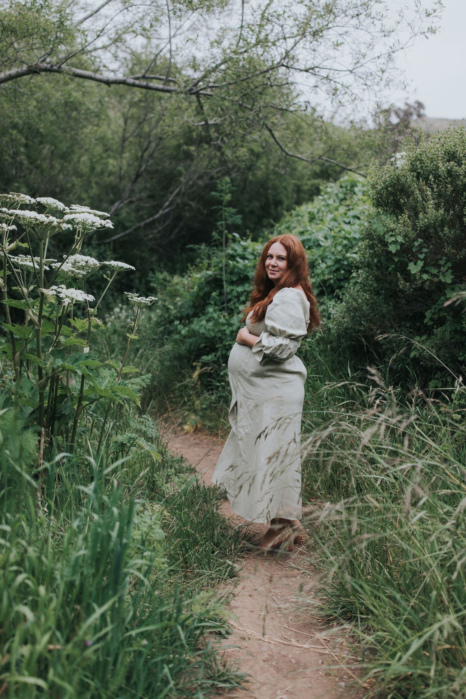 Expectant woman in flowy dress stands on nature trail among white flowering plants.