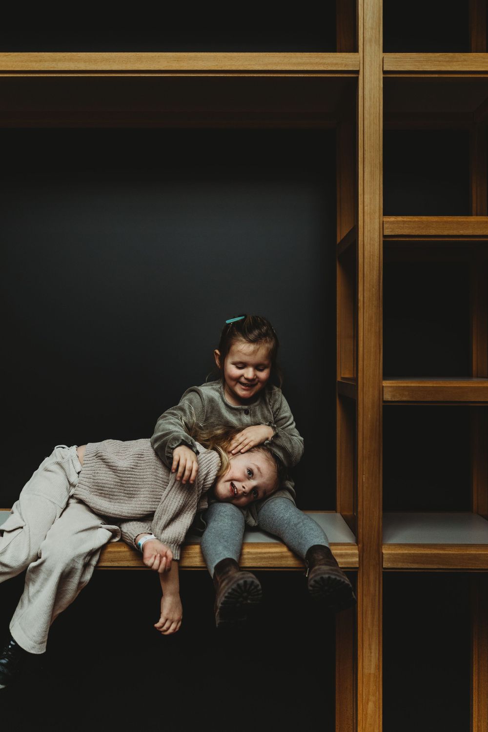 Two playful children rest on wooden shelving in a dark room.