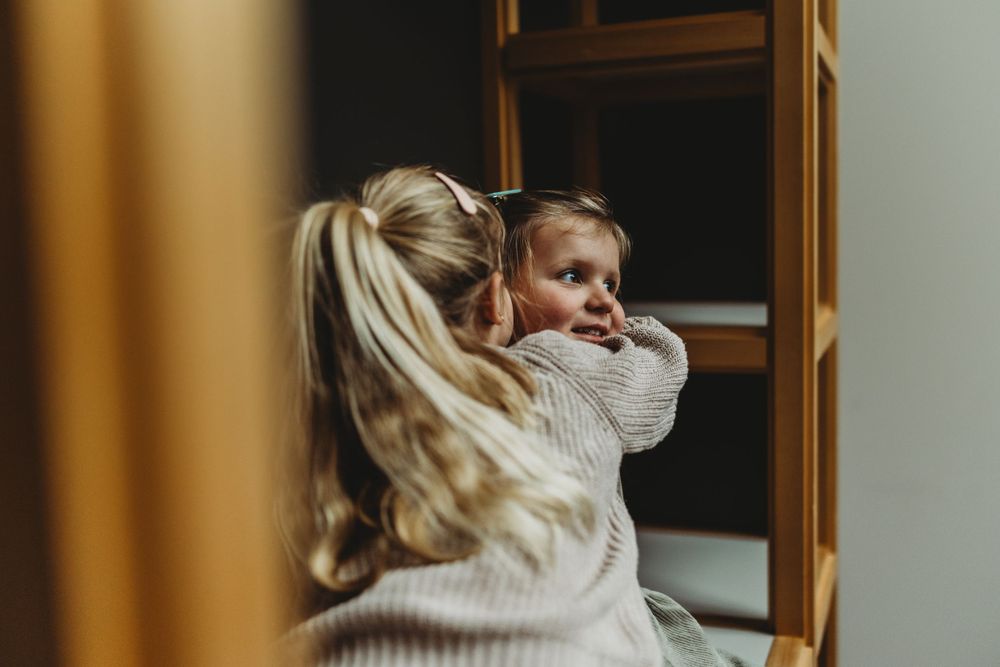 Two people in cozy sweaters share a warm embrace by a window in moody lighting.