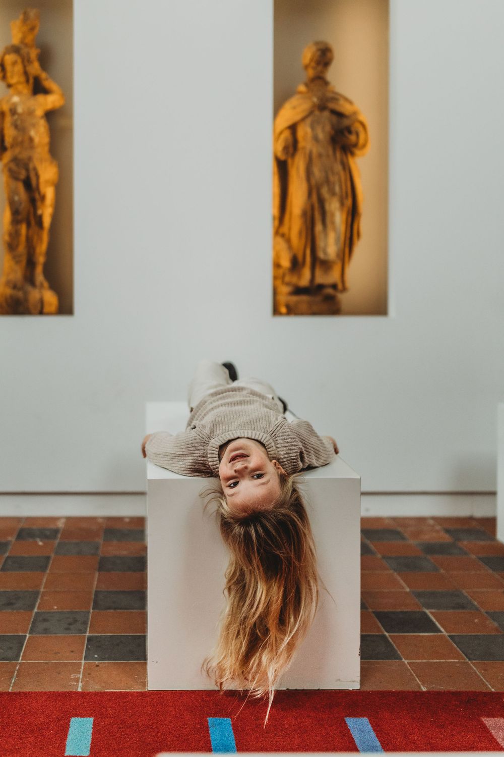 Person laying upside down on white pedestal with classical statues in background.