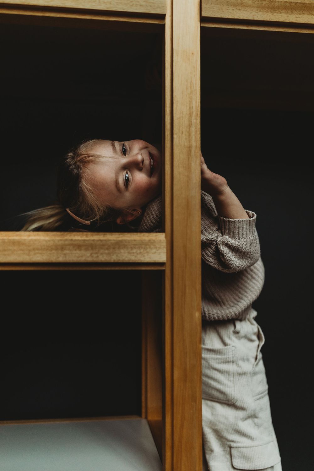 Child in gray sweater peeks through wooden shelving unit against dark background.