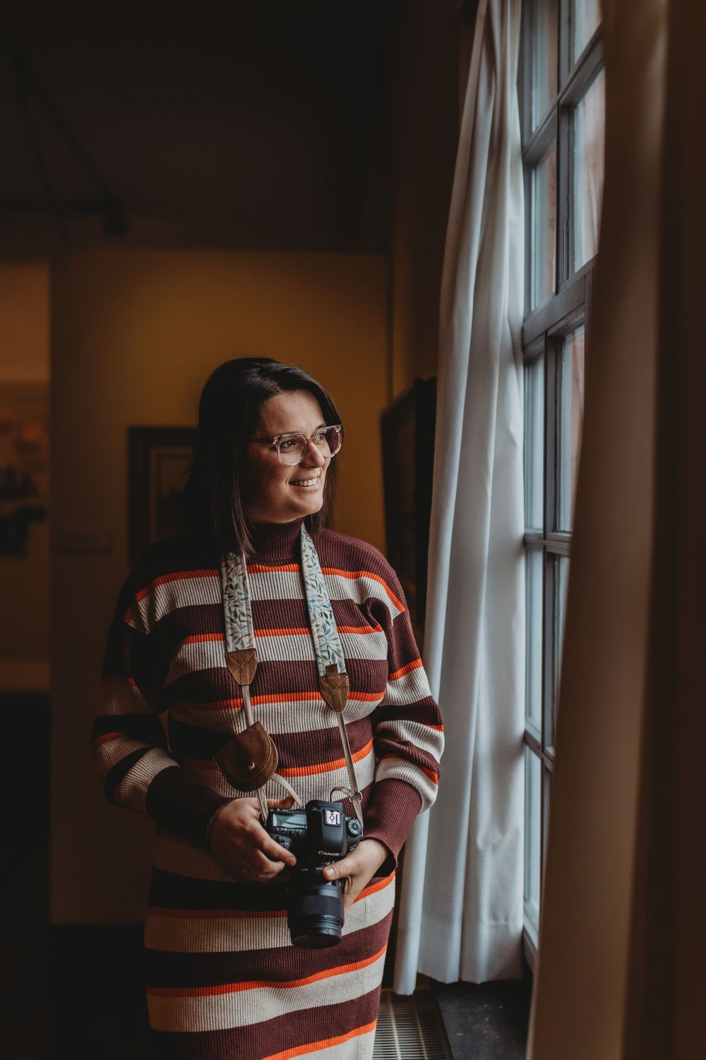 A photographer in a striped sweater dress stands by a window holding a vintage camera.