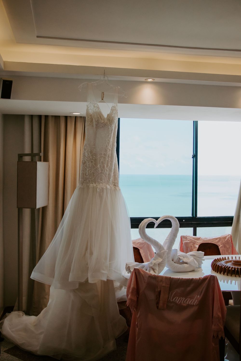 Wedding dress hanging in hotel room with ocean view and towel swans on table.