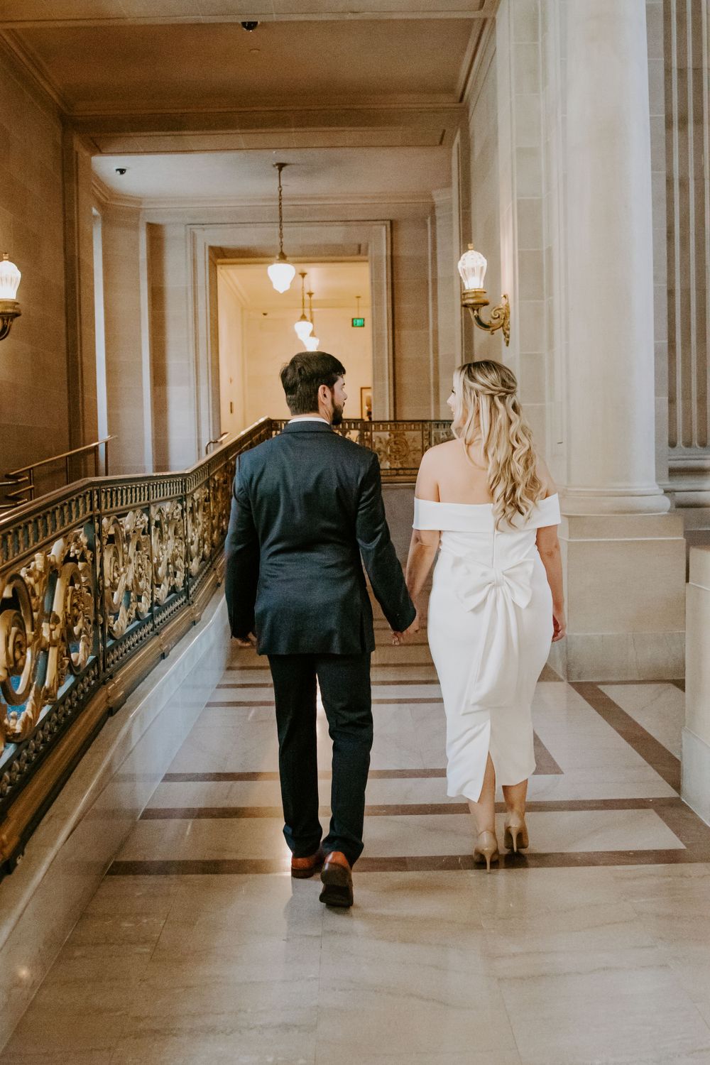 A couple walking hand in hand down an elegant hallway with marble columns and ornate railings.