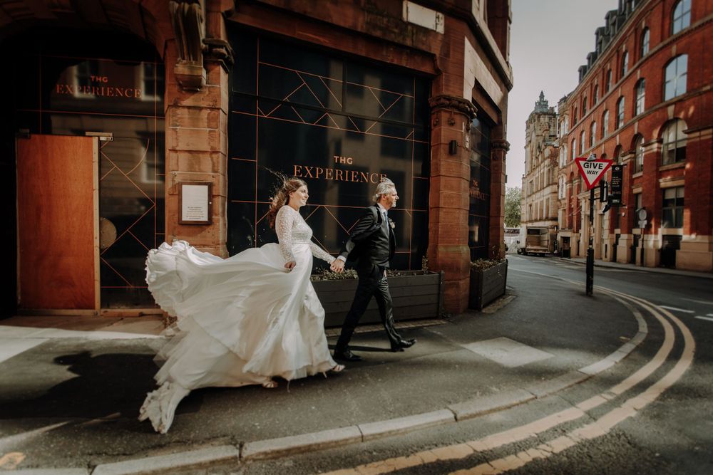 Bride's dress billows dramatically on city street corner with historic architecture backdrop.