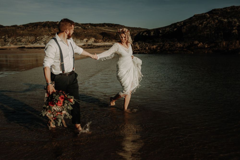 A couple holding hands and dancing at the water's edge with mountains in the background.