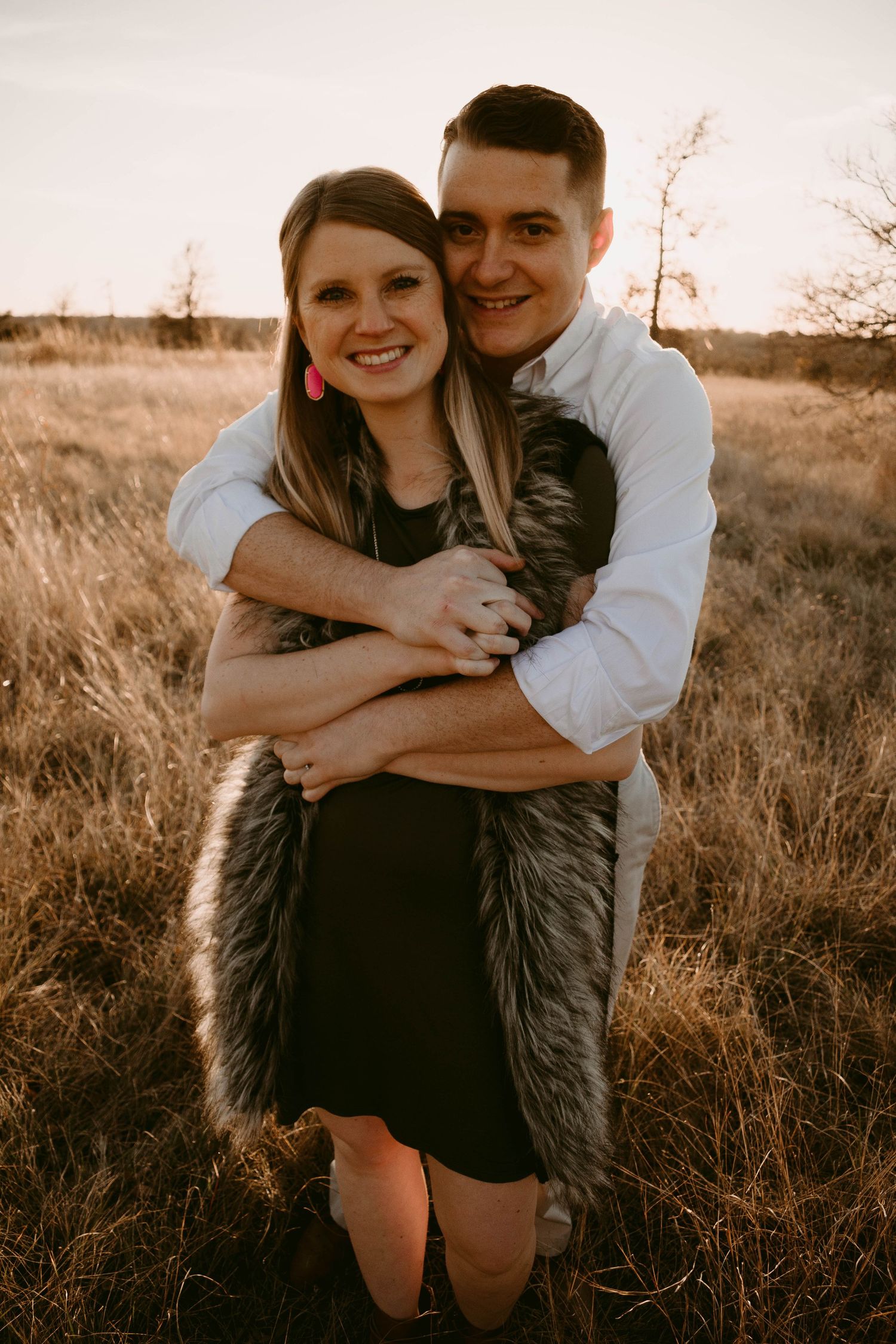 Happy couple sharing warm embrace while wearing fur vest in rustic field setting.