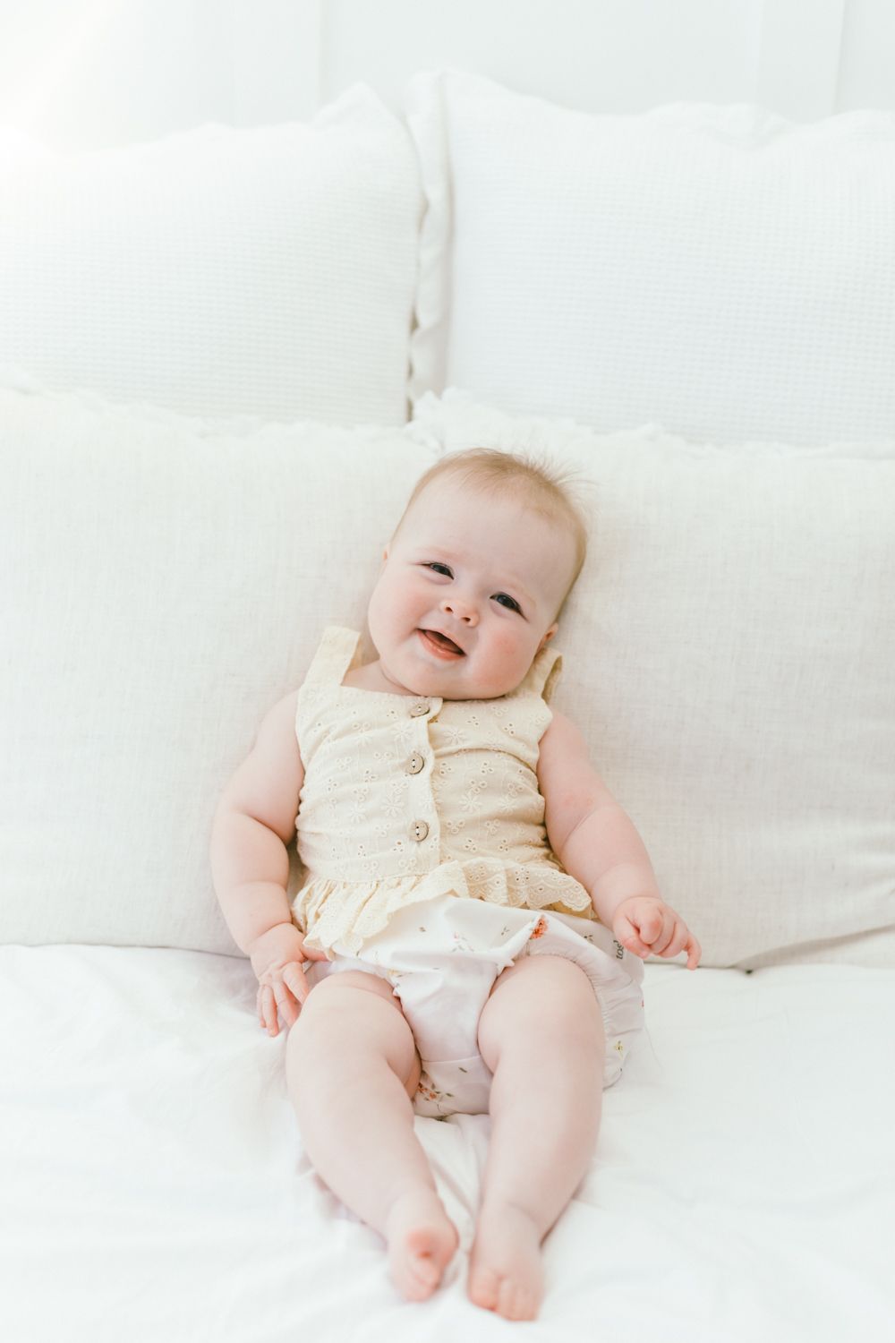 Baby wearing cream-colored outfit lying on white cushions with a happy expression.