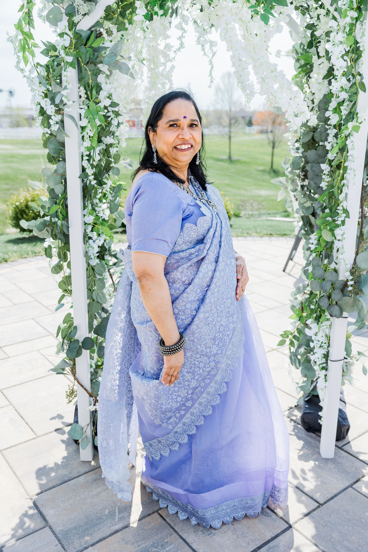 A woman in a light blue sari poses in an outdoor garden arch decorated with white flowers and greenery.