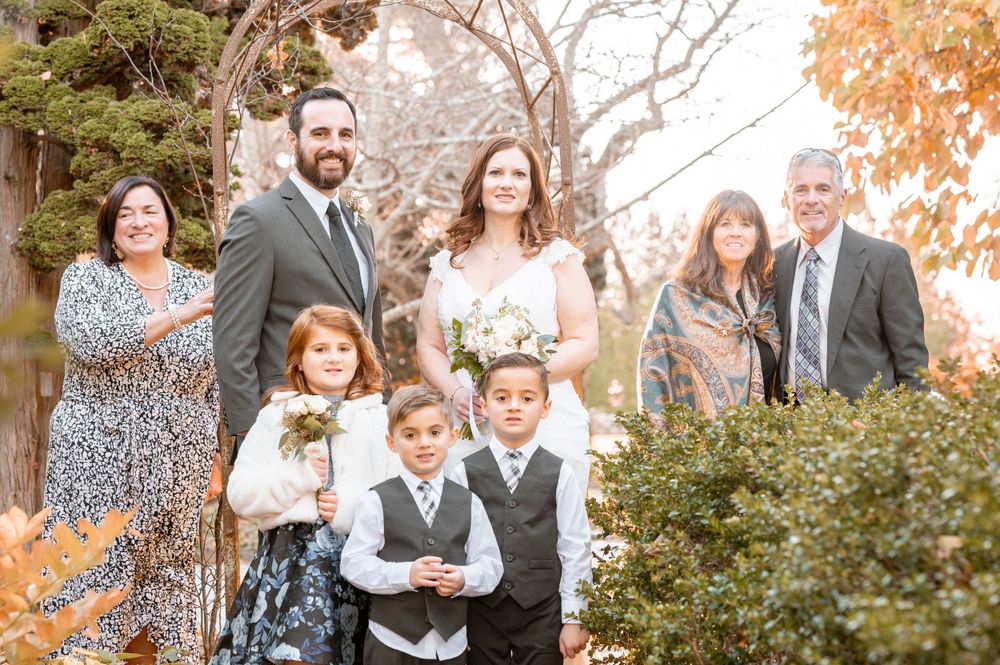 Wedding party gathers under garden arch surrounded by autumn foliage for formal portrait.