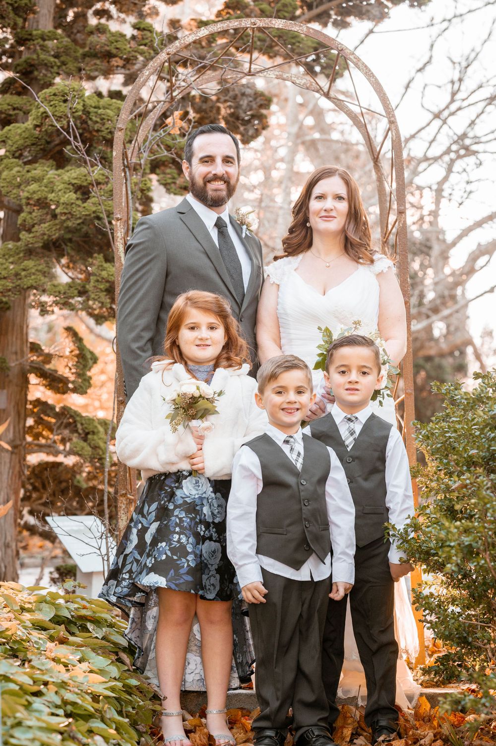Family group poses under rustic garden arch in formal wedding attire during autumn celebration.