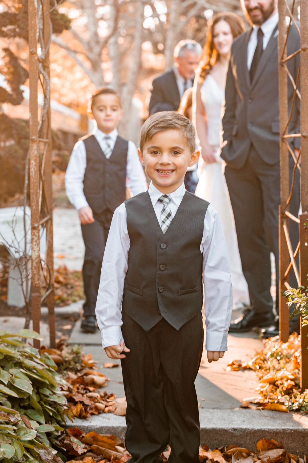 Young attendant in formal vest and tie stands smiling on garden path during autumn wedding ceremony.